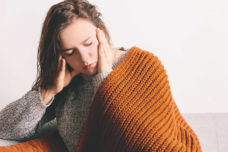 Rohrhof-Apotheke | Neuroinflammation und Mitochondropathie des Gehirns Eine Frau Sitze mit einer Wolldecke über den Schultern auf einer Couch und fasst mit beiden Händen ihre Schläfen an. Die Augen sind geschlossen. Sie ist erschöpft und scheint Schmerzen zu haben.