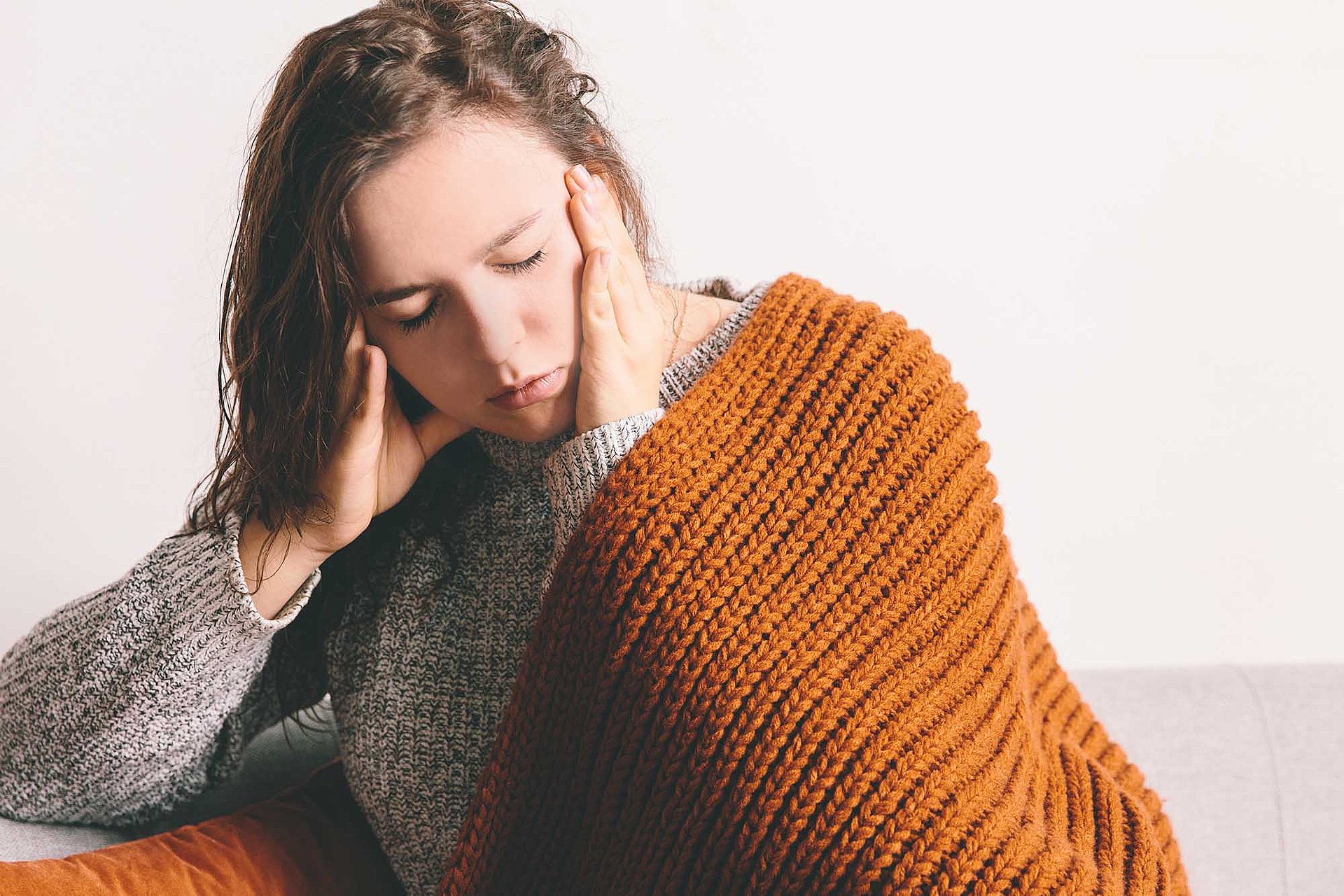 Rohrhof-Apotheke | Neuroinflammation und Mitochondropathie des Gehirns Eine Frau Sitze mit einer Wolldecke über den Schultern auf einer Couch und fasst mit beiden Händen ihre Schläfen an. Die Augen sind geschlossen. Sie ist erschöpft und scheint Schmerzen zu haben.