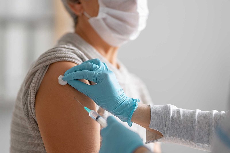 Rohrhof-Apotheke | Corona-Infektion/Impfung Eine Person mit Mundschutz und hochgekrempelten Ärmel bekommt den Arm vorbereitet für eine Impfung mit einer Spritze.
